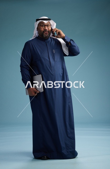 بورتريه لرجل عربي سعودي خليجي يرتدي ثوب كحلي وغتره بيضاء يستخدم الهاتف المحمول ويمسك جهاز التبليت باليد الاخرى, اجراء مكالمه هاتفيه, الجسم بالطول الكامل, خلفيه زرقاء