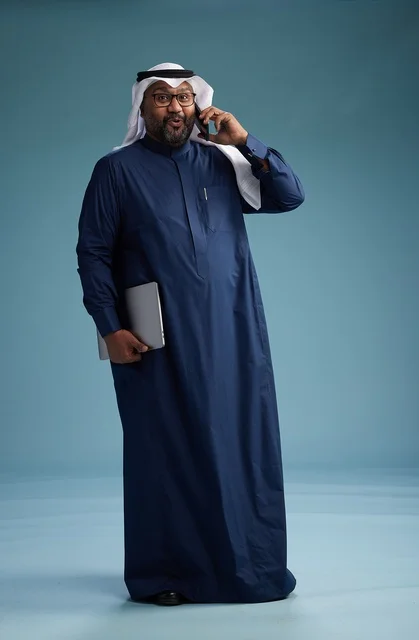 A portrait of an Arab Saudi Gulf man wearing a navy blue thobe and a white ghutrah, using a mobile phone and holding a tablet in the other hand, making a phone call, full body, blue background.