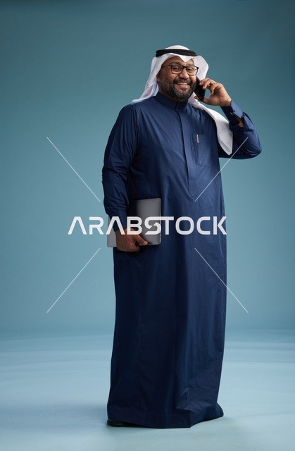 بورتريه لرجل عربي سعودي خليجي يرتدي ثوب كحلي وغتره بيضاء يستخدم الهاتف المحمول ويمسك جهاز التبليت باليد الاخرى, اجراء مكالمه هاتفيه, الجسم بالطول الكامل, خلفيه زرقاء