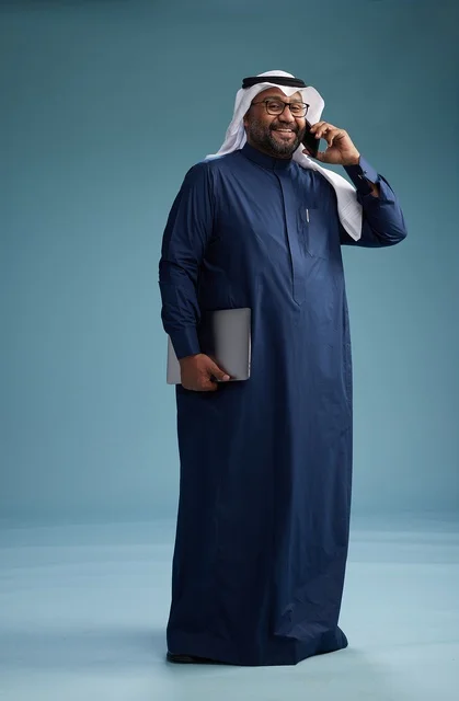A portrait of an Arab Saudi Gulf man wearing a navy blue thobe and a white ghutrah, using a mobile phone and holding a tablet in the other hand, making a phone call, full body, blue background.