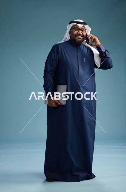 بورتريه لرجل عربي سعودي خليجي يرتدي ثوب كحلي وغتره بيضاء يستخدم الهاتف المحمول ويمسك جهاز التبليت باليد الاخرى, اجراء مكالمه هاتفيه, الجسم بالطول الكامل, خلفيه زرقاء