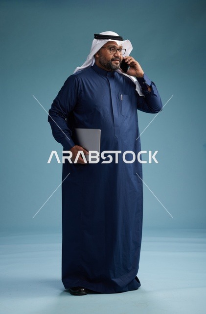 بورتريه لرجل عربي سعودي خليجي يرتدي ثوب كحلي وغتره بيضاء يستخدم الهاتف المحمول ويمسك جهاز التبليت باليد الاخرى, اجراء مكالمه هاتفيه, الجسم بالطول الكامل, خلفيه زرقاء