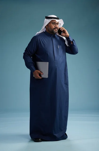 A portrait of an Arab Saudi Gulf man wearing a navy blue thobe and a white ghutrah, using a mobile phone and holding a tablet in the other hand, making a phone call, full body, blue background.