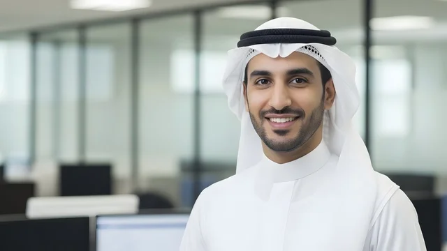 Looking at the camera with expressions of joy, business technology in the United Arab Emirates, a close-up shot of a smiling Emirati employee wearing the kandura and white ghutrah in a modern office, a professional work environment, the concept of perfection and focus.