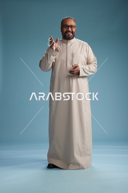 بورتريه لرجل عربي سعودي خليجي مبتسم يرتدي ثوب بيج يمسك بيده سبحه وينظر الى الكاميرا, كامل الجسم, خلفيه زرقاء