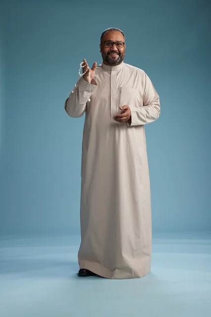 A portrait of a smiling Arab Saudi Gulf man wearing a beige thobe, holding a rosary in his hand and looking at the camera, full body, with a blue background.