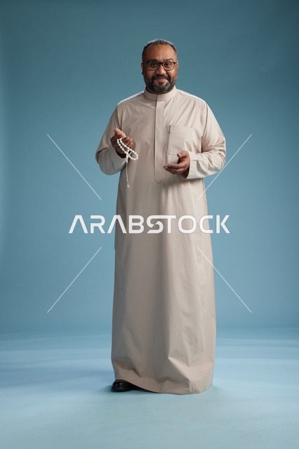 بورتريه لرجل عربي سعودي خليجي مبتسم يرتدي ثوب بيج يمسك بيده سبحه وينظر الى الكاميرا, كامل الجسم, خلفيه زرقاء