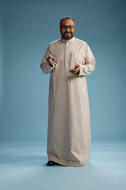 A portrait of a smiling Arab Saudi Gulf man wearing a beige thobe, holding a rosary in his hand and looking at the camera, full body, with a blue background.