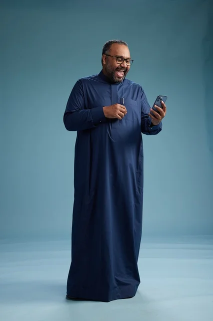 A portrait of a Saudi man wearing a navy thobe using a smartphone, communicating via mobile, full body, blue background.