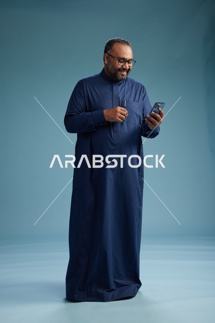 بورتريه لرجل سعودي يرتدي ثوب كحلي يستخدم الهاتف الذكي, التواصل عبر الجوال, الجسم بالكامل, خلفيه زرقاء