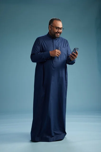 A portrait of a Saudi man wearing a navy thobe using a smartphone, communicating via mobile, full body, blue background.