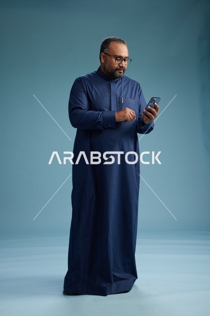 A portrait of a Saudi man wearing a navy thobe using a smartphone, communicating via mobile, full body, blue background.