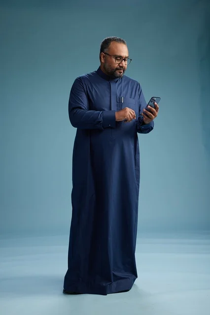 A portrait of a Saudi man wearing a navy thobe using a smartphone, communicating via mobile, full body, blue background.