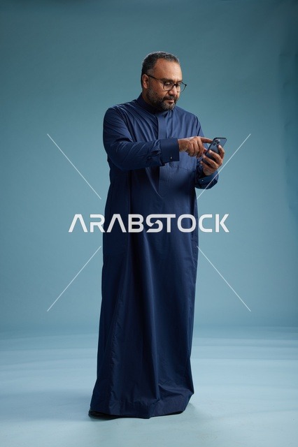 A portrait of a Saudi man wearing a navy thobe using a smartphone, communicating via mobile, full body, blue background.