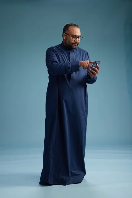 A portrait of a Saudi man wearing a navy thobe using a smartphone, communicating via mobile, full body, blue background.