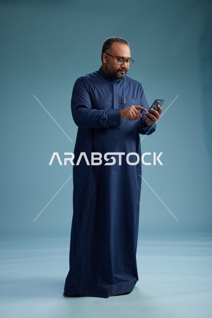 بورتريه لرجل سعودي يرتدي ثوب كحلي يستخدم الهاتف الذكي, التواصل عبر الجوال, الجسم بالكامل, خلفيه زرقاء