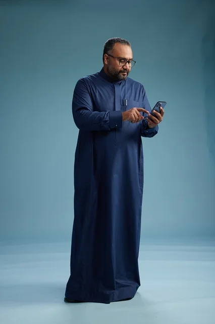 A portrait of a Saudi man wearing a navy thobe using a smartphone, communicating via mobile, full body, blue background.