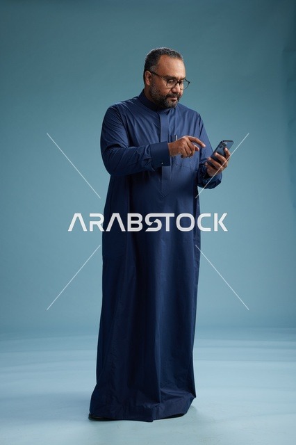 A portrait of a Saudi man wearing a navy thobe using a smartphone, communicating via mobile, full body, blue background.
