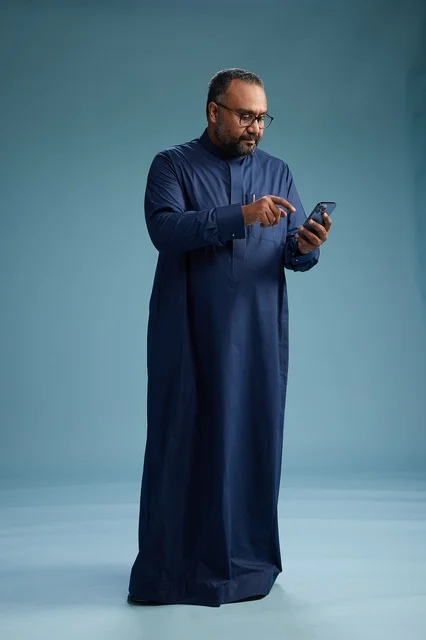 بورتريه لرجل سعودي يرتدي ثوب كحلي يستخدم الهاتف الذكي, التواصل عبر الجوال, الجسم بالكامل, خلفيه زرقاء