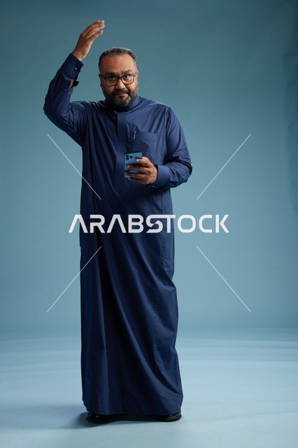 بورتريه لرجل سعودي يرتدي ثوب كحلي يستخدم الهاتف الذكي, التواصل عبر الجوال, الجسم بالكامل, خلفيه زرقاء