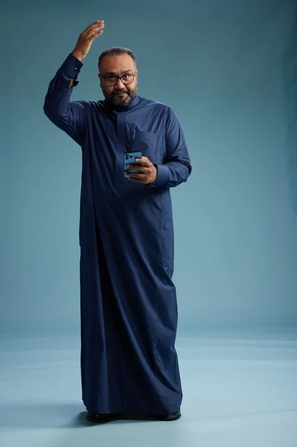 A portrait of a Saudi man wearing a navy thobe using a smartphone, communicating via mobile, full body, blue background.
