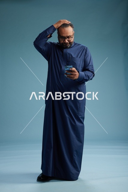 بورتريه لرجل سعودي يرتدي ثوب كحلي يستخدم الهاتف الذكي, التواصل عبر الجوال, الجسم بالكامل, خلفيه زرقاء