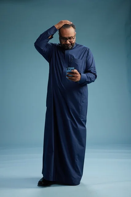 A portrait of a Saudi man wearing a navy thobe using a smartphone, communicating via mobile, full body, blue background.