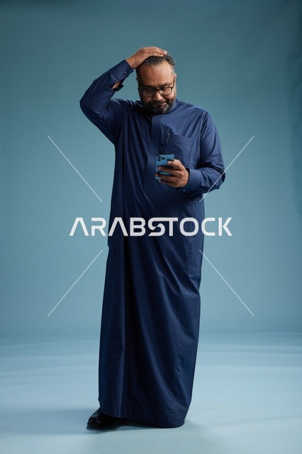 A portrait of a Saudi man wearing a navy thobe using a smartphone, communicating via mobile, full body, blue background.
