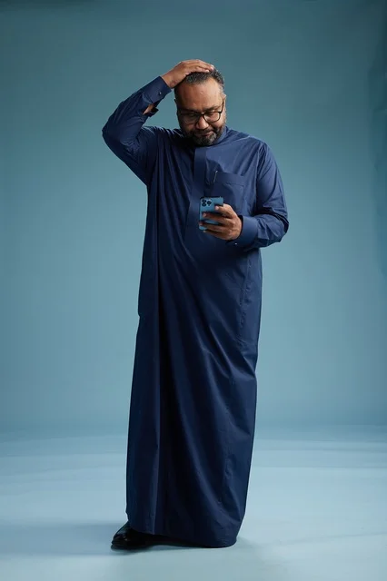 A portrait of a Saudi man wearing a navy thobe using a smartphone, communicating via mobile, full body, blue background. A portrait of a Saudi man wearing a navy thobe using a smartphone, communicating via mobile, full body, blue background.