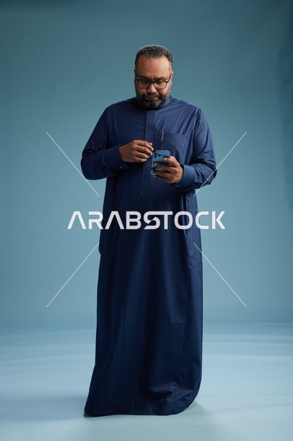 بورتريه لرجل سعودي يرتدي ثوب كحلي يستخدم الهاتف الذكي, التواصل عبر الجوال, الجسم بالكامل, خلفيه زرقاء