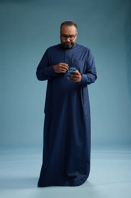A portrait of a Saudi man wearing a navy thobe using a smartphone, communicating via mobile, full body, blue background.
