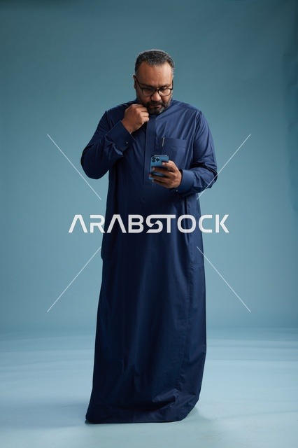 A portrait of a Saudi man wearing a navy thobe using a smartphone, communicating via mobile, full body, blue background.