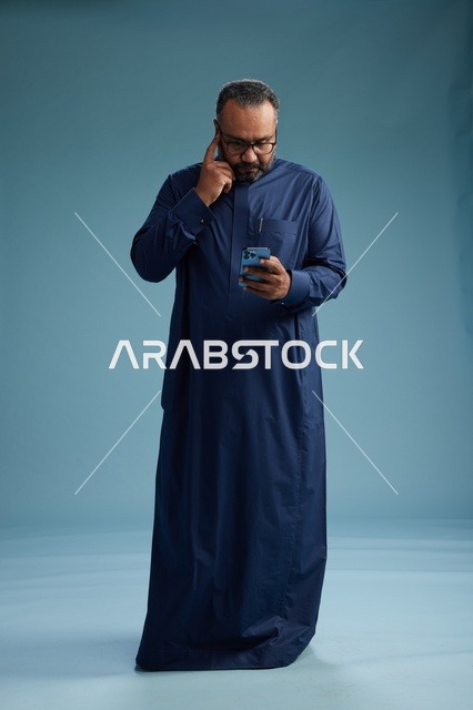 A portrait of a Saudi man wearing a navy thobe using a smartphone, communicating via mobile, full body, blue background.