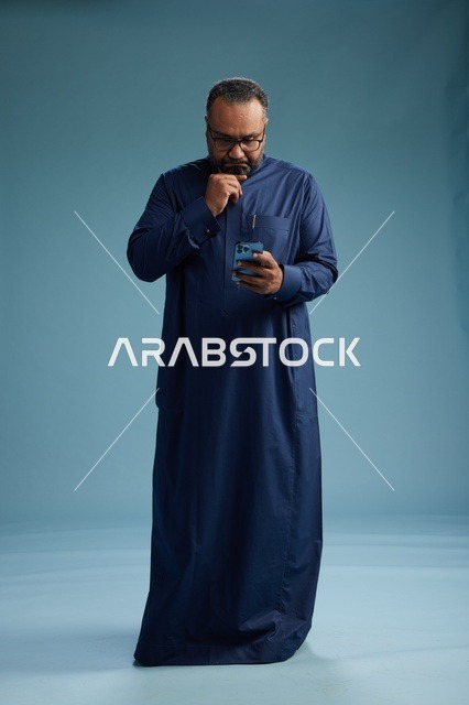 بورتريه لرجل سعودي يرتدي ثوب كحلي يستخدم الهاتف الذكي, التواصل عبر الجوال, الجسم بالكامل, خلفيه زرقاء