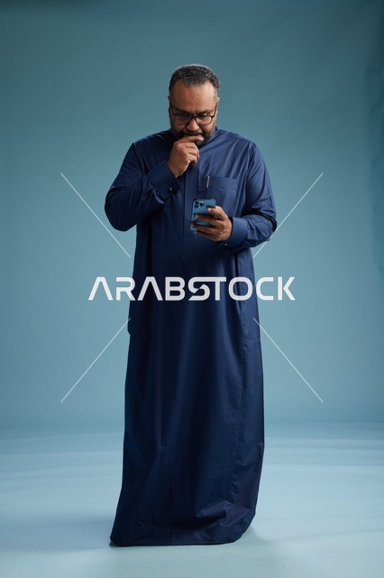 بورتريه لرجل سعودي يرتدي ثوب كحلي يستخدم الهاتف الذكي, التواصل عبر الجوال, الجسم بالكامل, خلفيه زرقاء