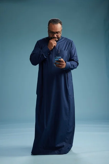 A portrait of a Saudi man wearing a navy thobe using a smartphone, communicating via mobile, full body, blue background.