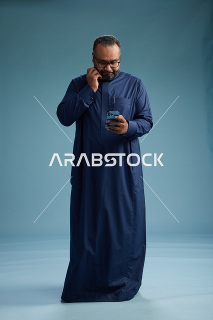 بورتريه لرجل سعودي يرتدي ثوب كحلي يستخدم الهاتف الذكي, التواصل عبر الجوال, الجسم بالكامل, خلفيه زرقاء