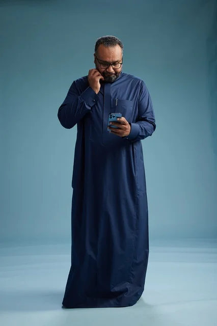 A portrait of a Saudi man wearing a navy thobe using a smartphone, communicating via mobile, full body, blue background.