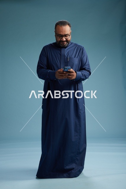 بورتريه لرجل سعودي يرتدي ثوب كحلي يستخدم الهاتف الذكي, التواصل عبر الجوال, الجسم بالكامل, خلفيه زرقاء