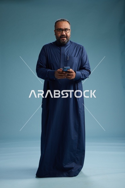 بورتريه لرجل سعودي يرتدي ثوب كحلي يستخدم الهاتف الذكي, التواصل عبر الجوال, الجسم بالكامل, خلفيه زرقاء