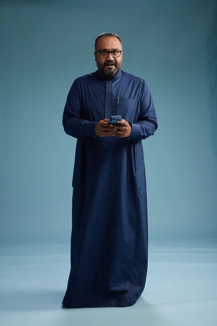 A portrait of a Saudi man wearing a navy thobe using a smartphone, communicating via mobile, full body, blue background.