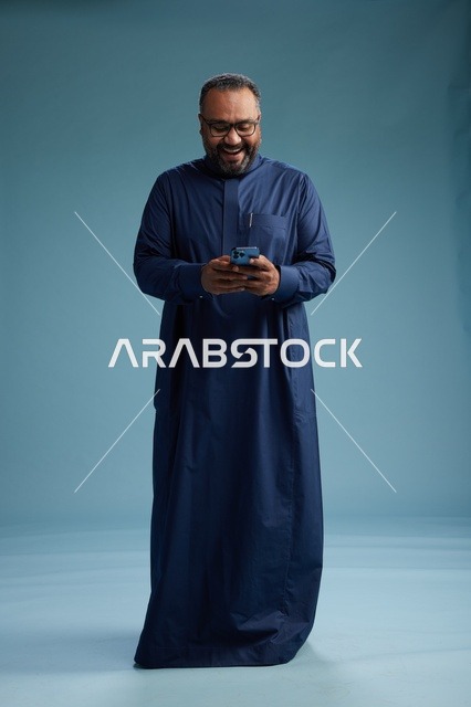 بورتريه لرجل سعودي يرتدي ثوب كحلي يستخدم الهاتف الذكي, التواصل عبر الجوال, الجسم بالكامل, خلفيه زرقاء