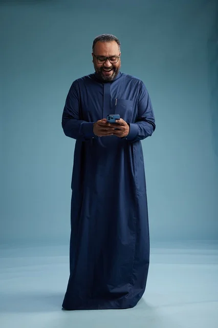 A portrait of a Saudi man wearing a navy thobe using a smartphone, communicating via mobile, full body, blue background.