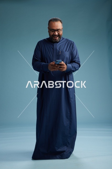بورتريه لرجل سعودي يرتدي ثوب كحلي يستخدم الهاتف الذكي, التواصل عبر الجوال, الجسم بالكامل, خلفيه زرقاء