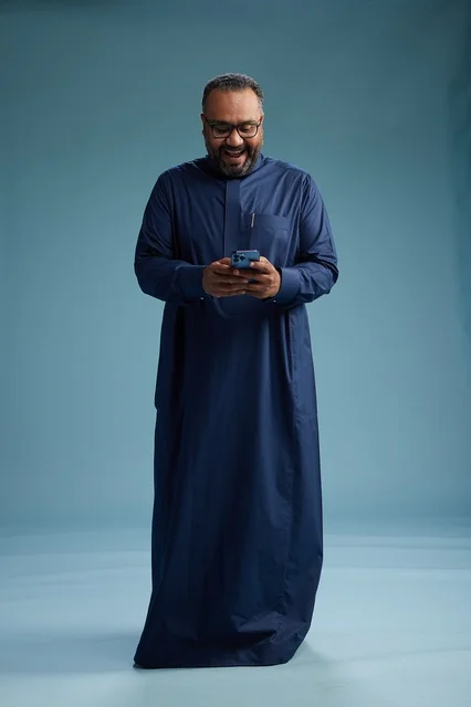 A portrait of a Saudi man wearing a navy thobe using a smartphone, communicating via mobile, full body, blue background.