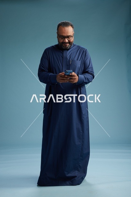 بورتريه لرجل سعودي يرتدي ثوب كحلي يستخدم الهاتف الذكي, التواصل عبر الجوال, الجسم بالكامل, خلفيه زرقاء