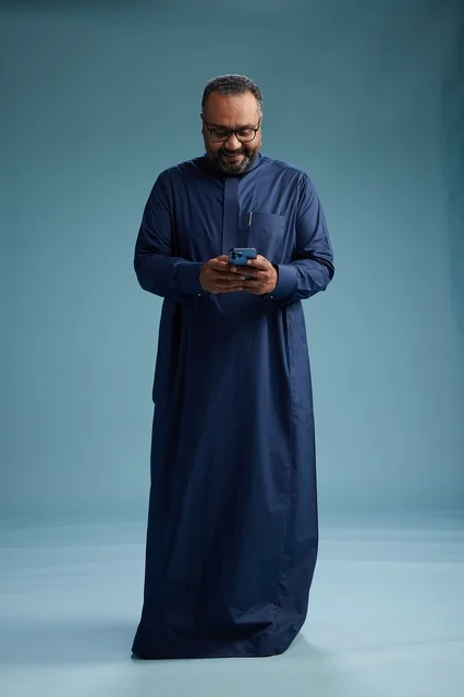 A portrait of a Saudi man wearing a navy thobe using a smartphone, communicating via mobile, full body, blue background.