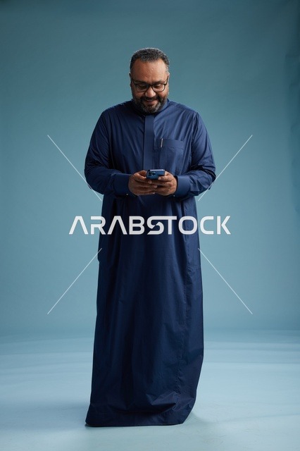 A portrait of a Saudi man wearing a navy thobe using a smartphone, communicating via mobile, full body, blue background.