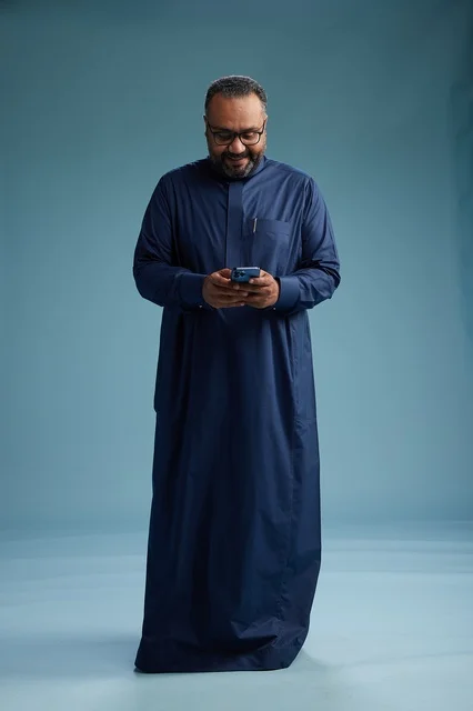 A portrait of a Saudi man wearing a navy thobe using a smartphone, communicating via mobile, full body, blue background.