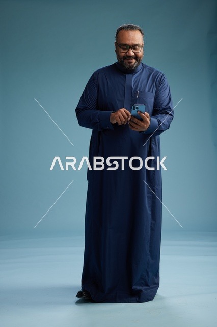 A portrait of a Saudi man wearing a navy thobe using a smartphone, communicating via mobile, full body, blue background.